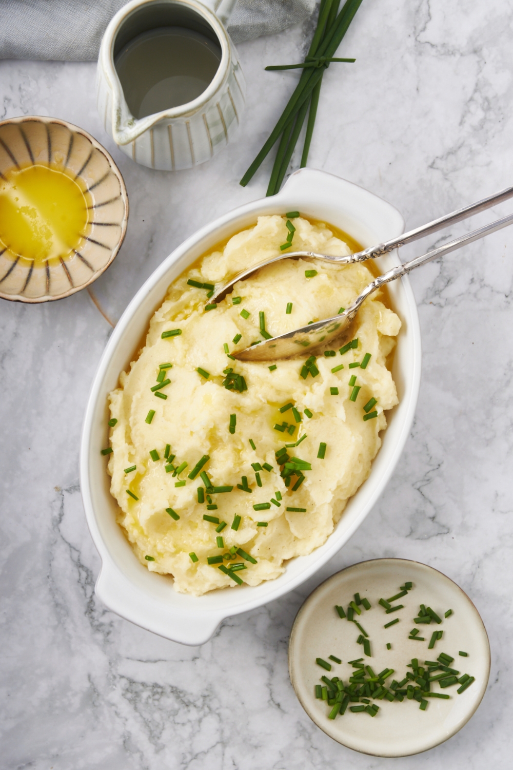 A white baking dish with mashed potatoes, two serving spoons, and fresh chives garnished on top. A plate of chives and a bowl of melted butter surrounds the baking dish.