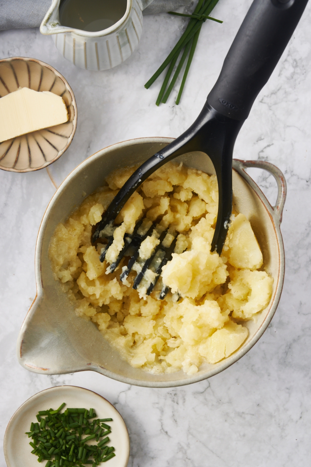 A mixing bowl with potatoes being mashed with a potato masher.