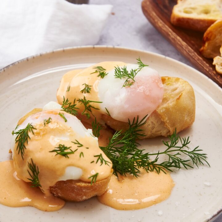 Two pieces of toast on a plate, each topped with a poached egg, Hollandaise sauce, and fresh dill.