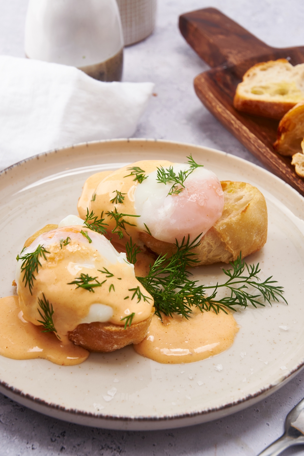 Two pieces of toast on a plate, each topped with a poached egg, Hollandaise sauce, and fresh dill.