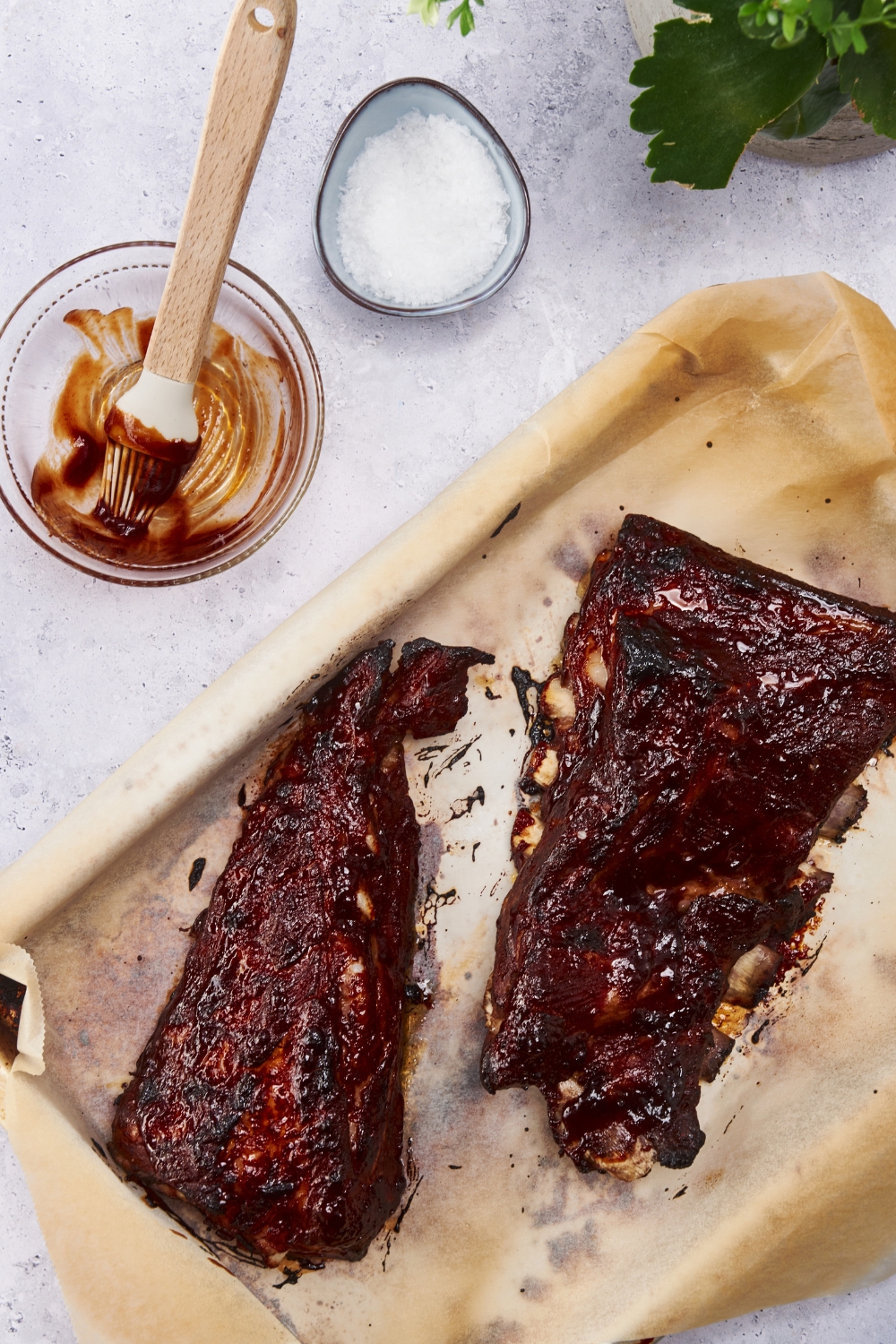 Overhead view of baked ribs covered in barbecue sauce. The ribs are on a baking sheet lined with parchment paper and there is an empty bowl of barbecue sauce with a basting brush next to the baking sheet.