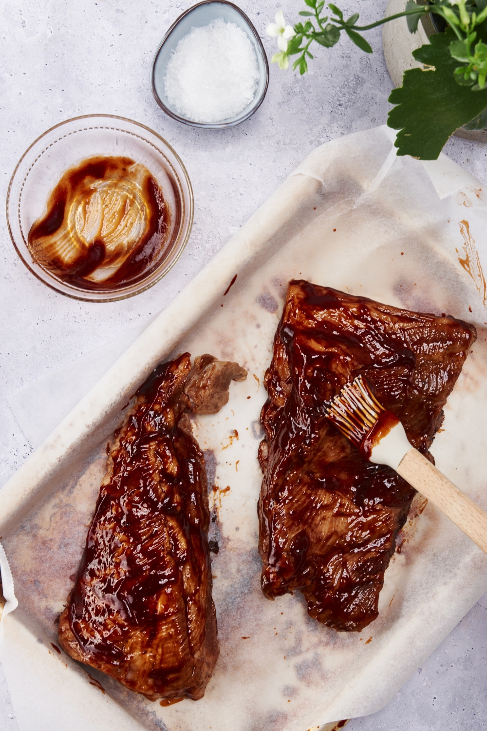 Cooked ribs being coated in barbecue sauce with a silicone basting brush. The ribs are on a baking sheet lined with parchment paper and there is an empty bowl of barbecue sauce next to the ribs.