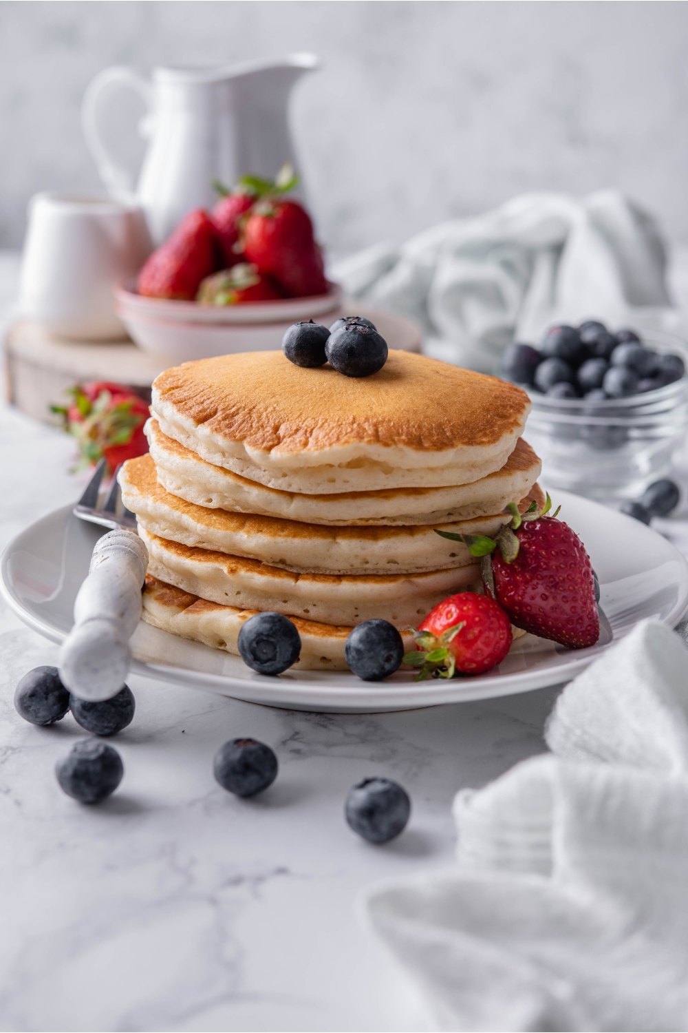A stack of pancakes topped with blueberries on a white plate with a fork and strawberries on the plate beside the pancakes. Berries are scattered on the counter and in the background.