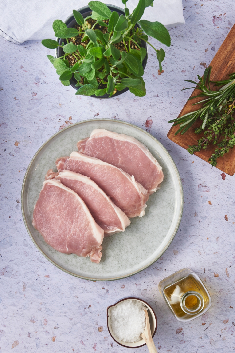 Four raw pork chops layered on top of each other surrounded by fresh herbs, a container of oil, and a bowl of salt.