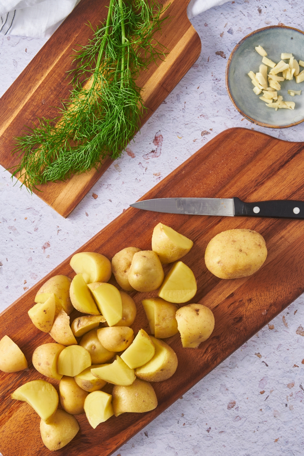 Overhead view of sliced potatoes on a cutting board with a knife on the cutting board and fresh dill on a second cutting board.