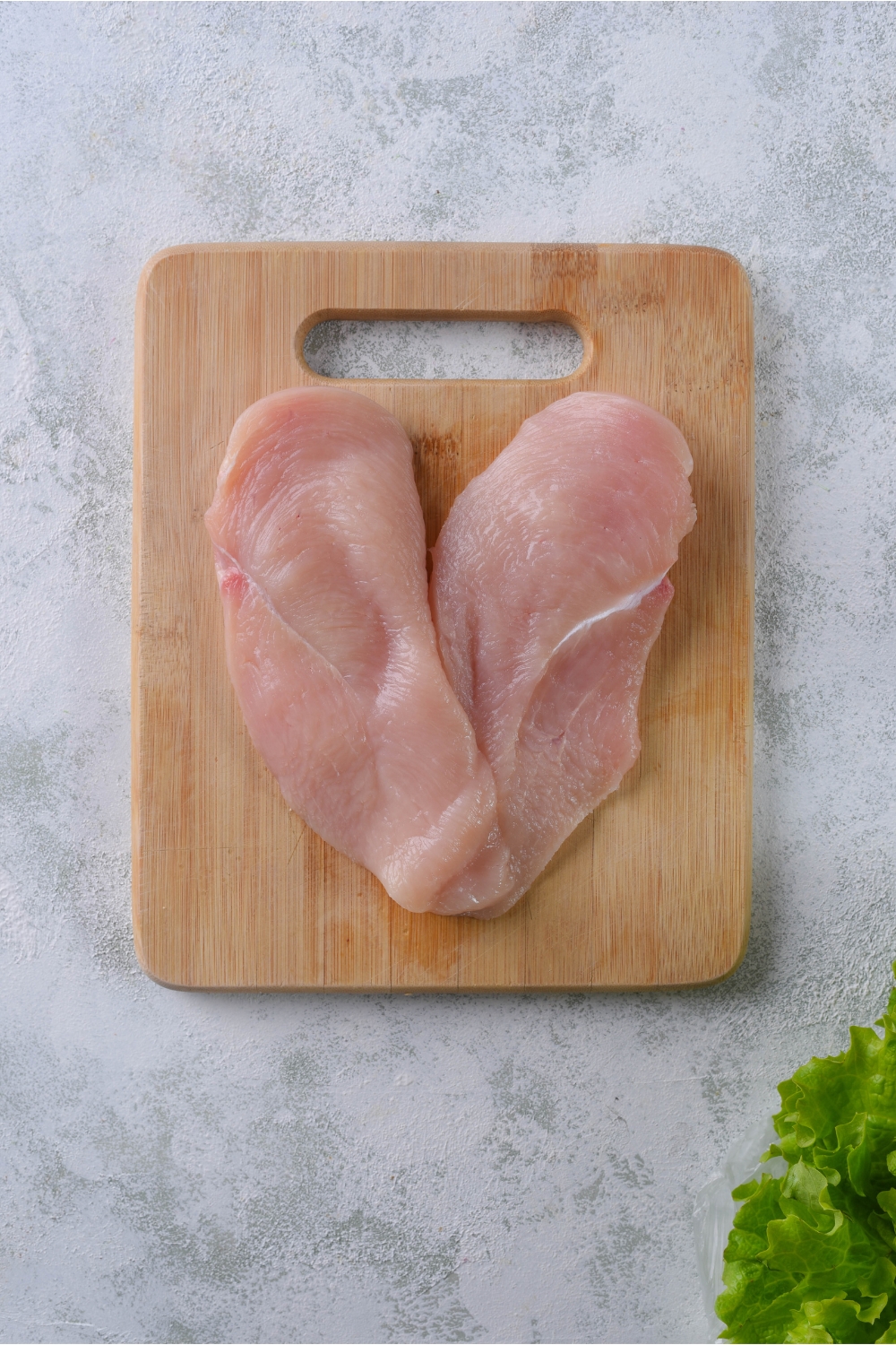 A wood cutting board with two raw chicken breast cutlets on it.