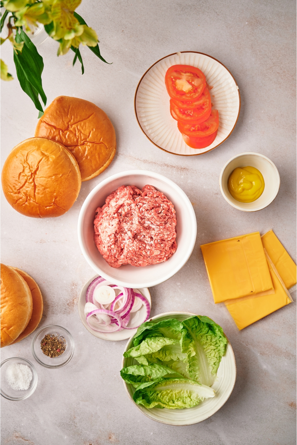 An assortment of ingredients including a bowl of raw ground beef, two buns, three slices of cheese, and plates of lettuce, red onion, sliced tomatoes, and mustard.