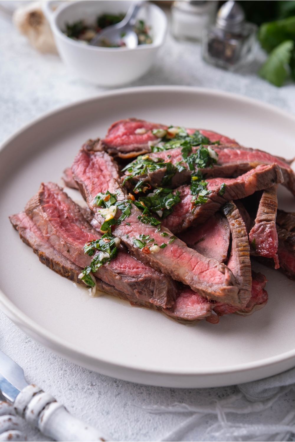 Thinly sliced bavette steak piled on a white plate and covered in chimichurri sauce.