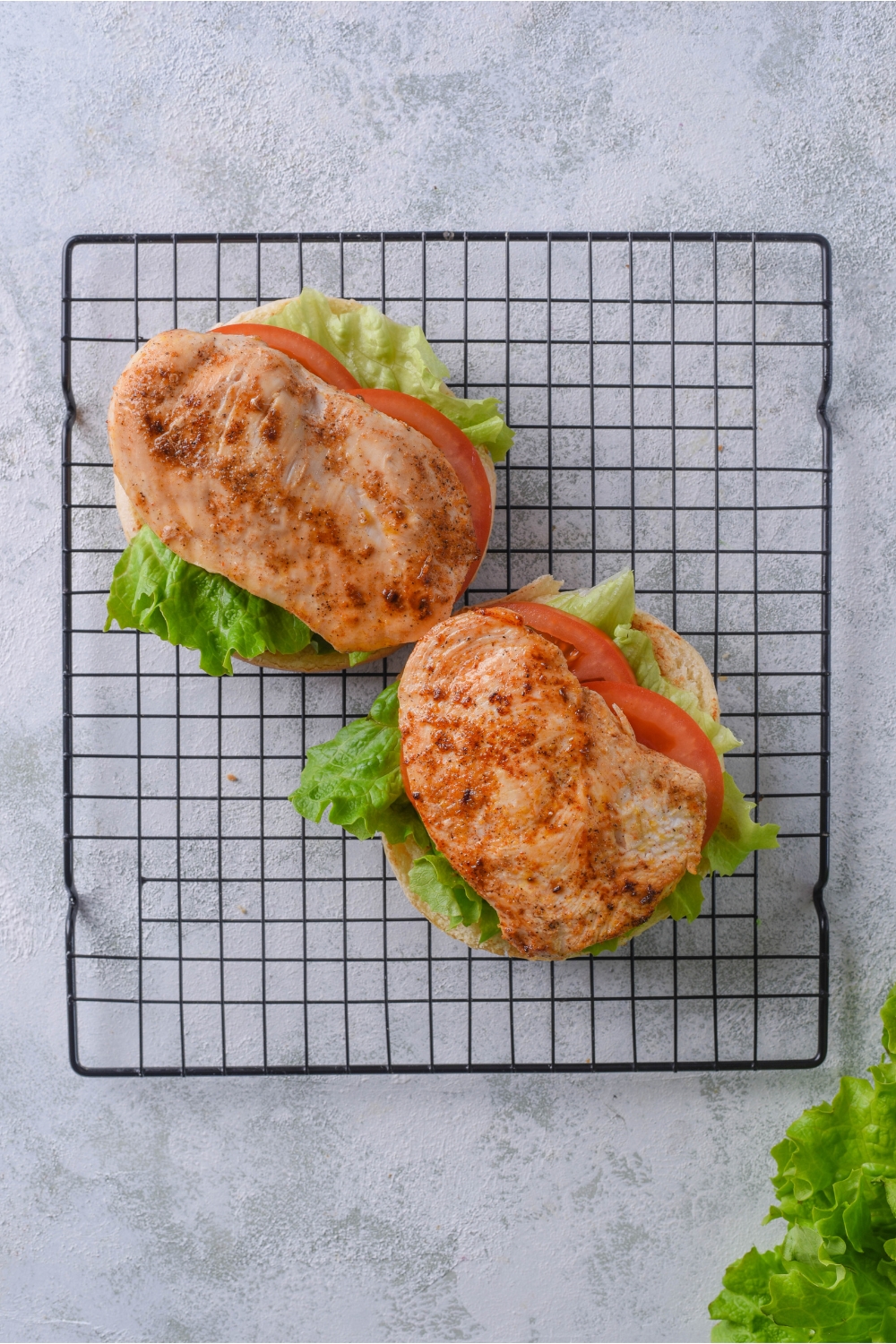 A wire rack with two hamburger buns, each topped with lettuce, two slices of tomato, and a slice of grilled chicken.