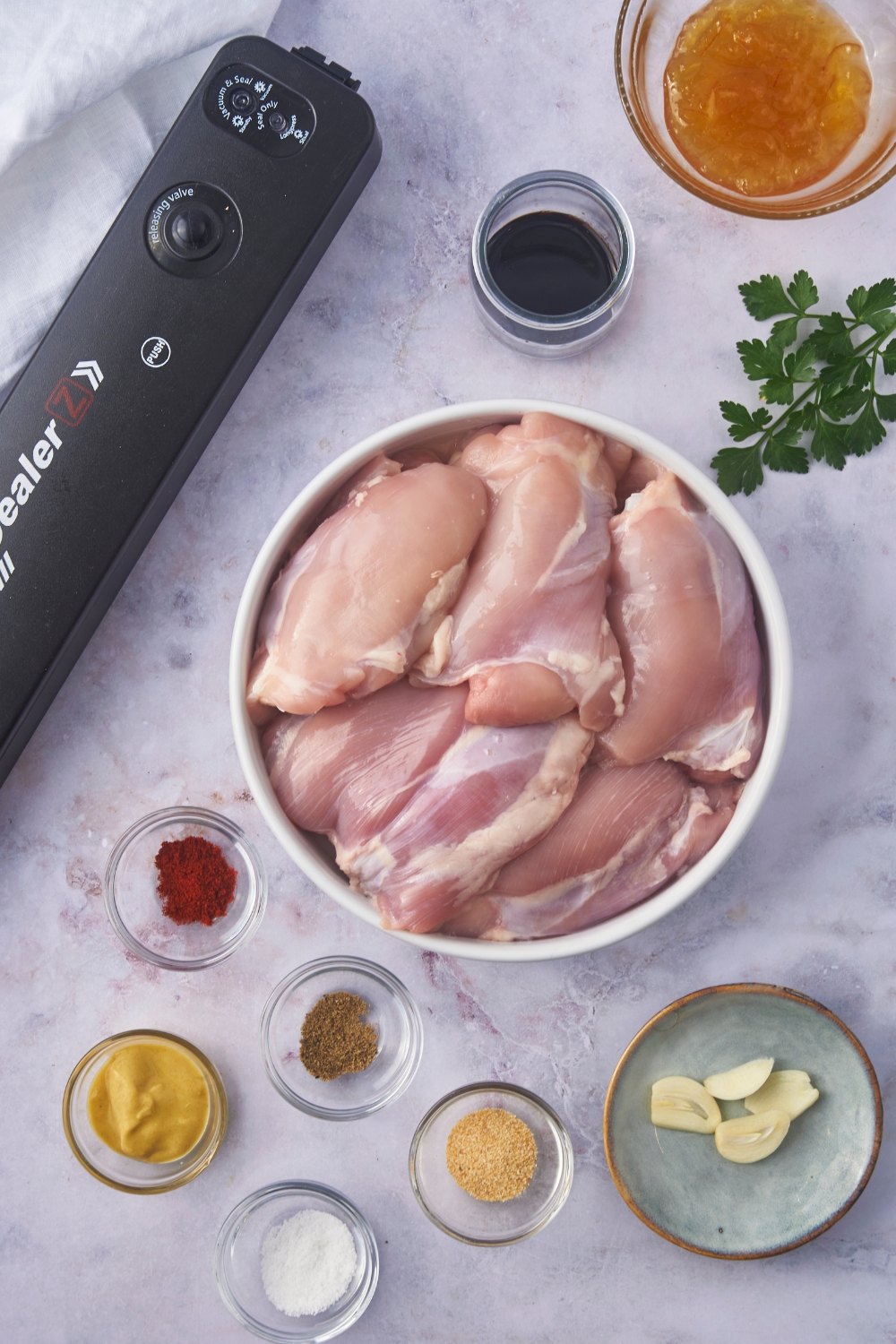 A vacuum-sealer next to an assortment of ingredients including a bowl of raw chicken thighs and small bowls of mustard, paprika, garlic, and orange marmalade.