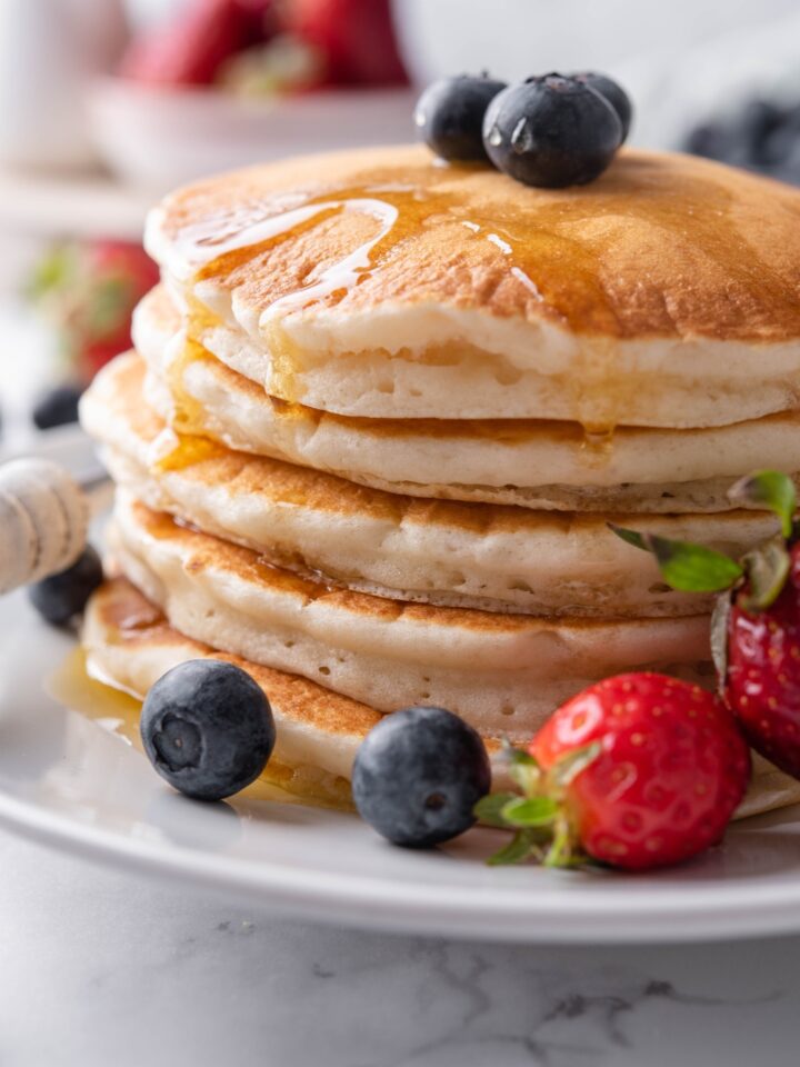 Close up of a stack of pancakes covered in maple syrup with berries on top of the pancakes and surrounding them on the plate.