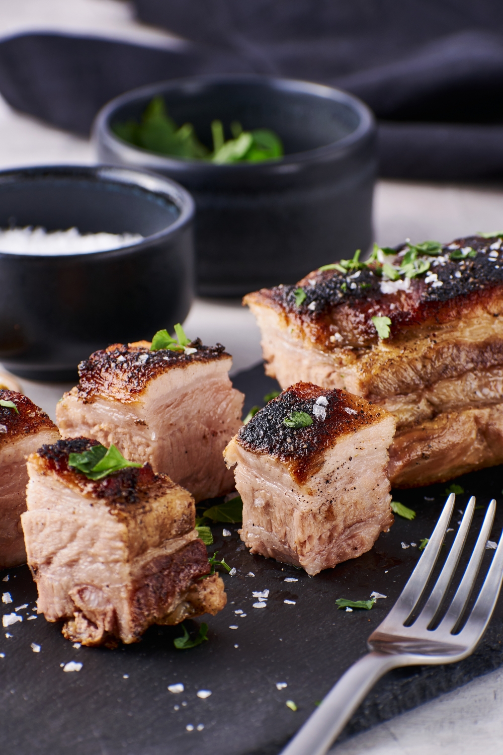 Cooked pork belly on a piece of black slate with a fork next to the pork. The pork is garnished with fresh herbs and salt.