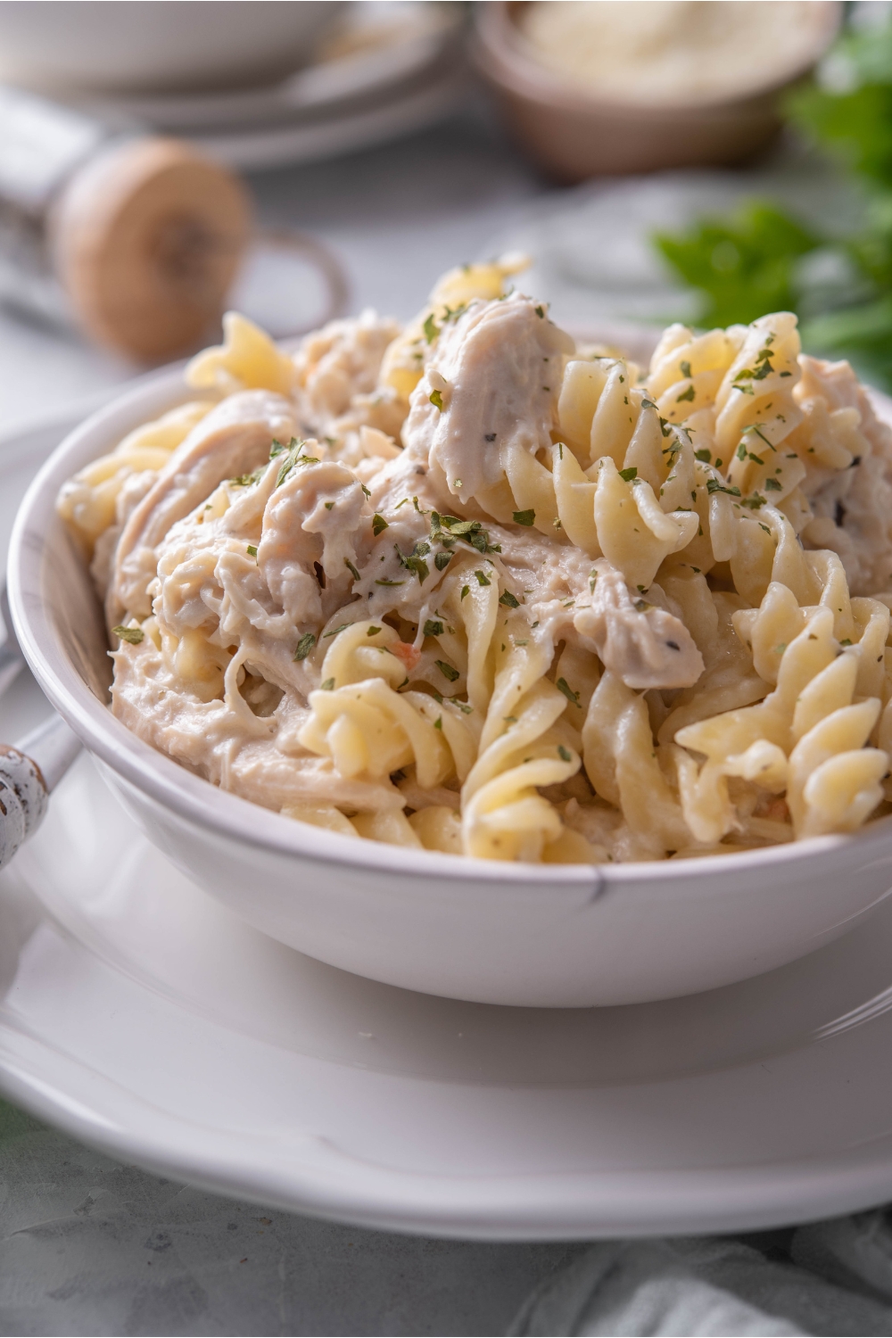 Close up of a bowl of pasta and shredded chicken garnished with fresh herbs. The bowl is on a white plate.