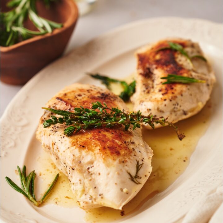 Two cooked chicken breasts on a white plate, each garnished with fresh herbs and covered in a butter sauce.