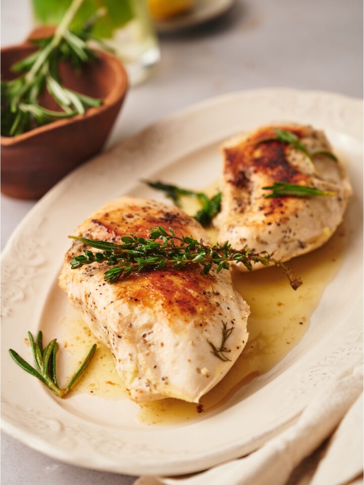 Two cooked chicken breasts on a white plate, each garnished with fresh herbs and covered in a butter sauce.