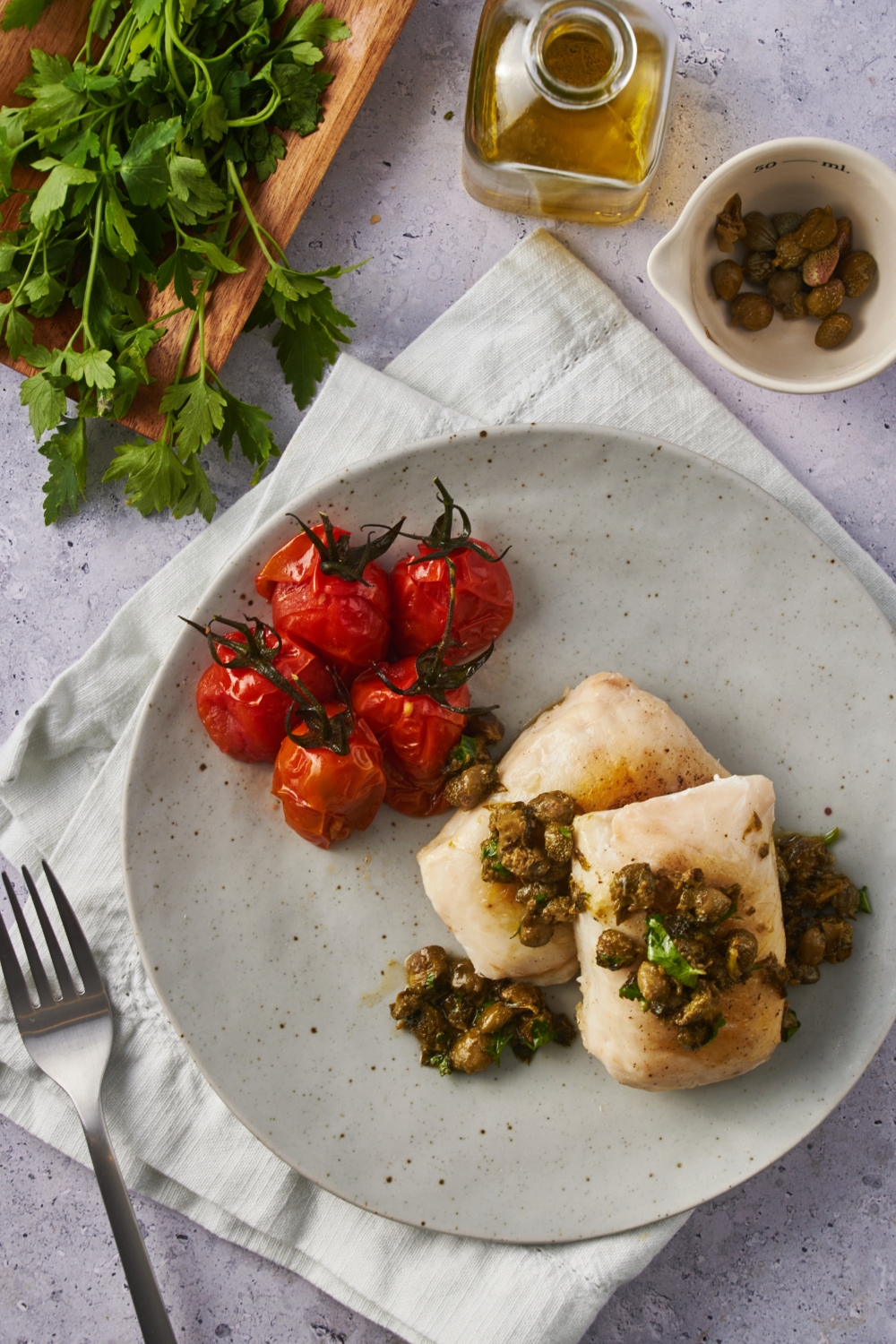 Overhead view of two cooked halibut fillets, both covered in a caper and herb mixture. Next to the fish are five roasted cherry tomatoes.