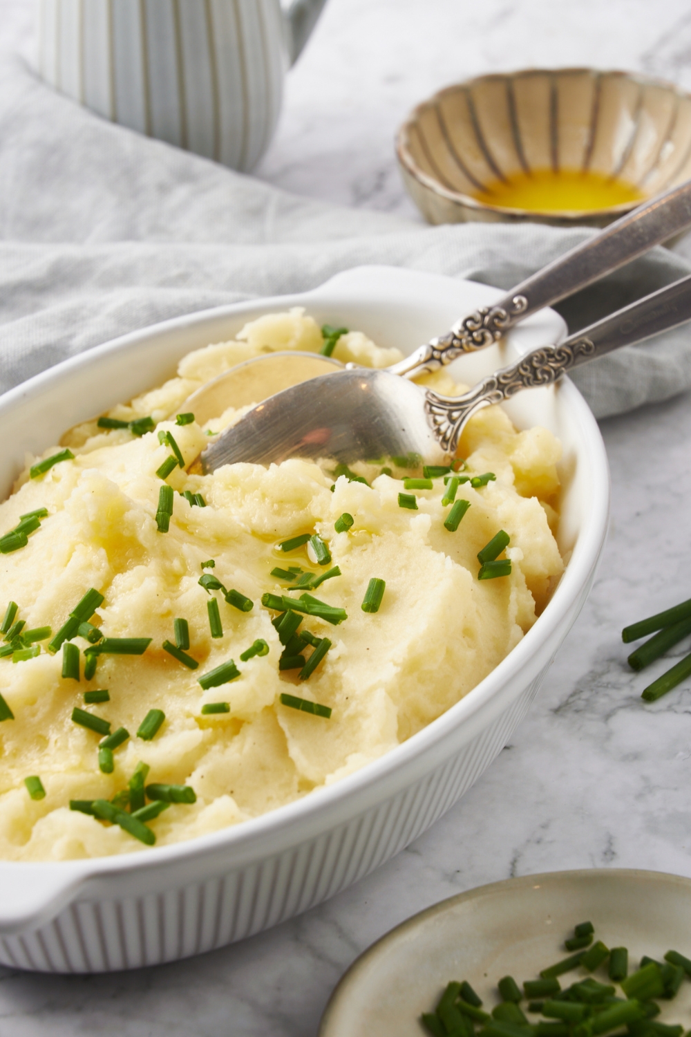 A white baking dish filled with mashed potatoes, two serving spoons, and fresh chives garnished on top.