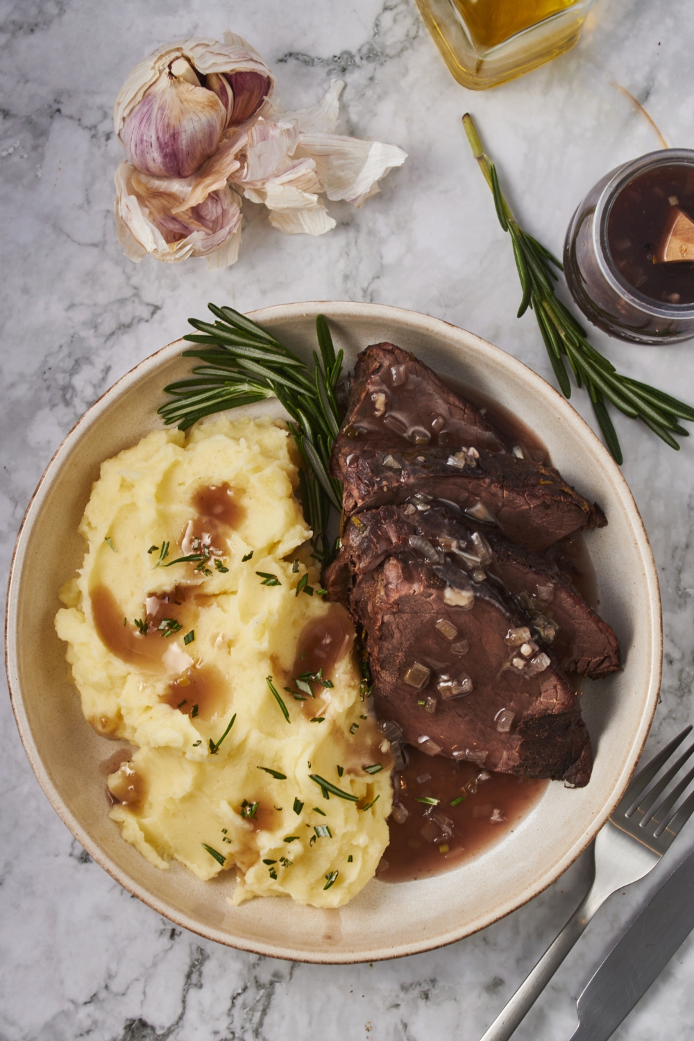 Overhead view of a plate of sliced chuck roast covered in a gravy with a fresh sprig of rosemary and a pile of mashed potatoes garnished with fresh herbs and some gravy poured over top of the potatoes.