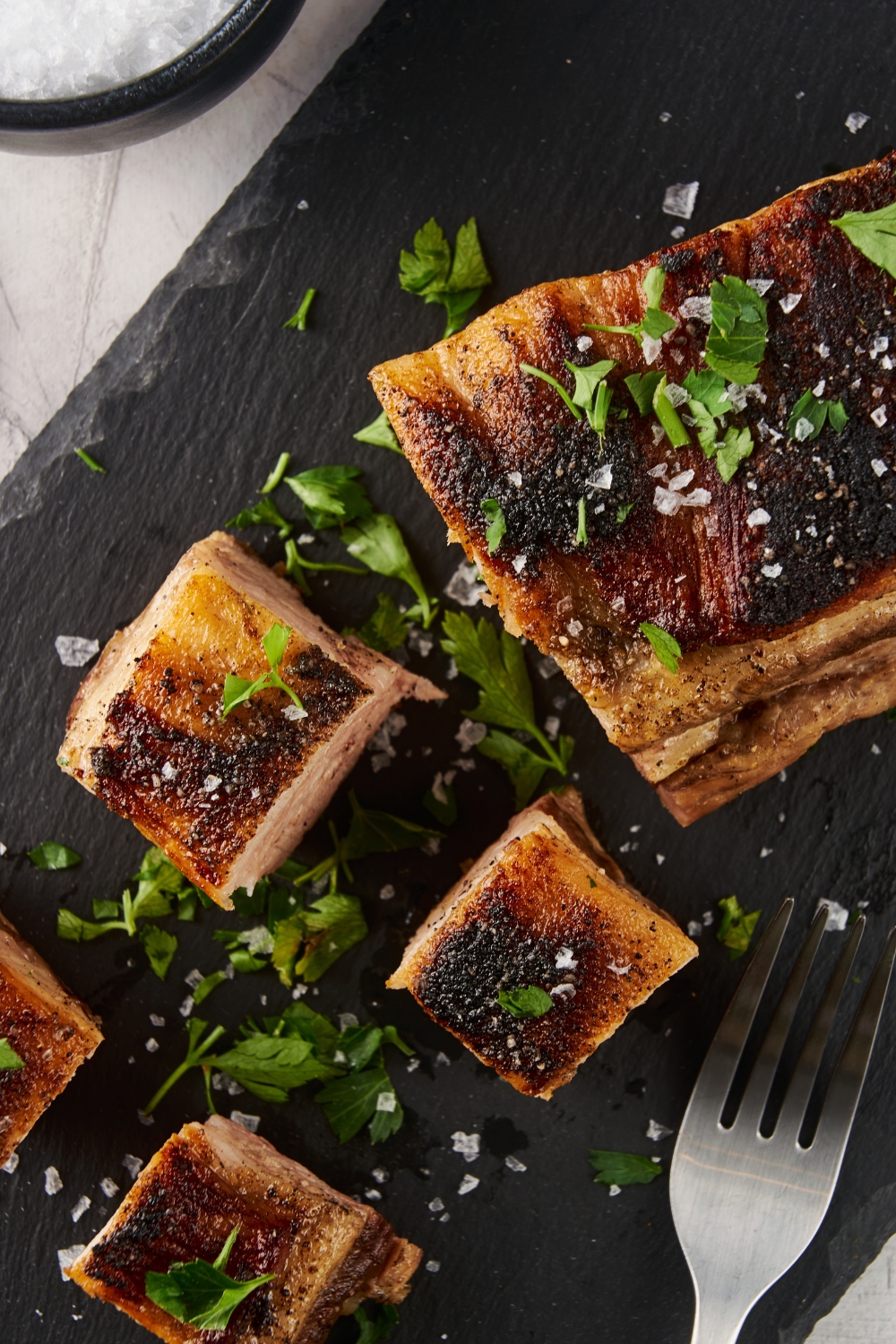 Overhead view of seared pork belly cut into squares. The pork belly is on a black piece of slate and is garnished with fresh parsley and coarse salt. There is a fork next to the pork belly.