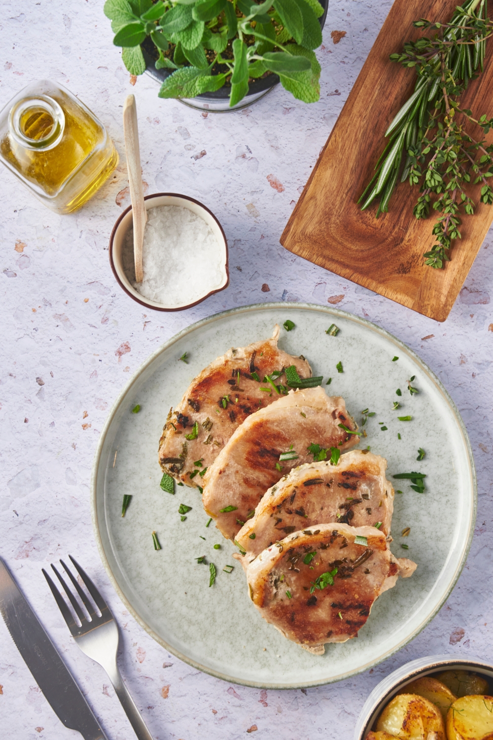 A blue plate with four seared pork chops layered on top of each other and garnished with fresh herbs. Surrounding the plate of pork chops is a bowl of salt, a container of oil, a fork and knife, and a wood tray with fresh herbs.