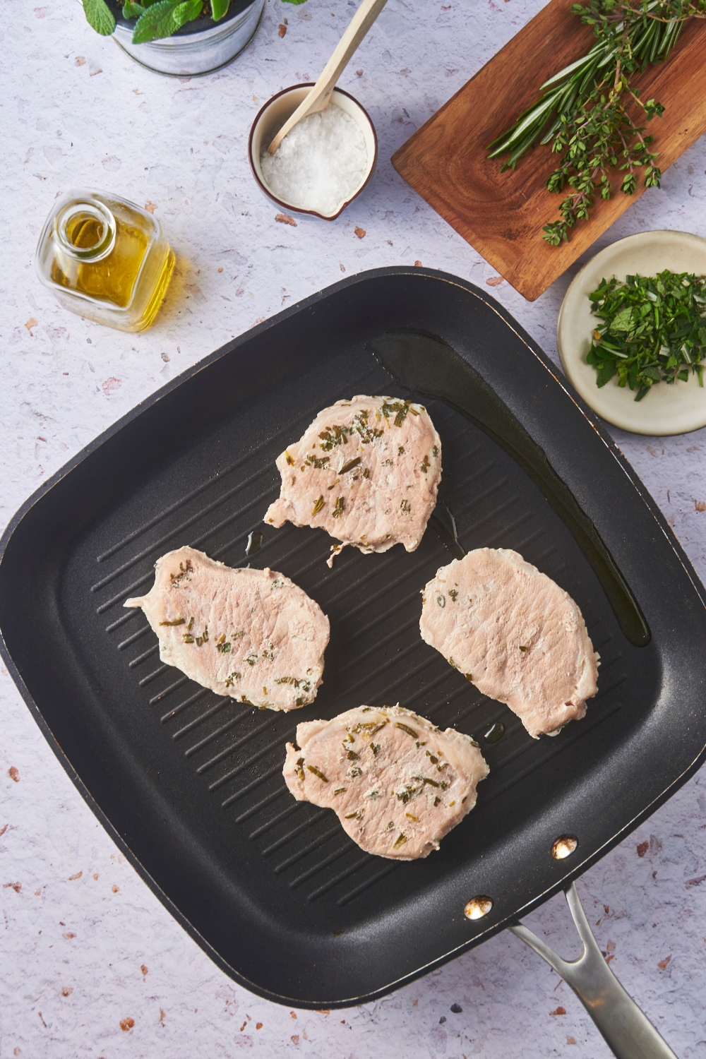 Four cooked pork chops on a black skillet, surrounded by ingredients including oil, salt, and fresh herbs.