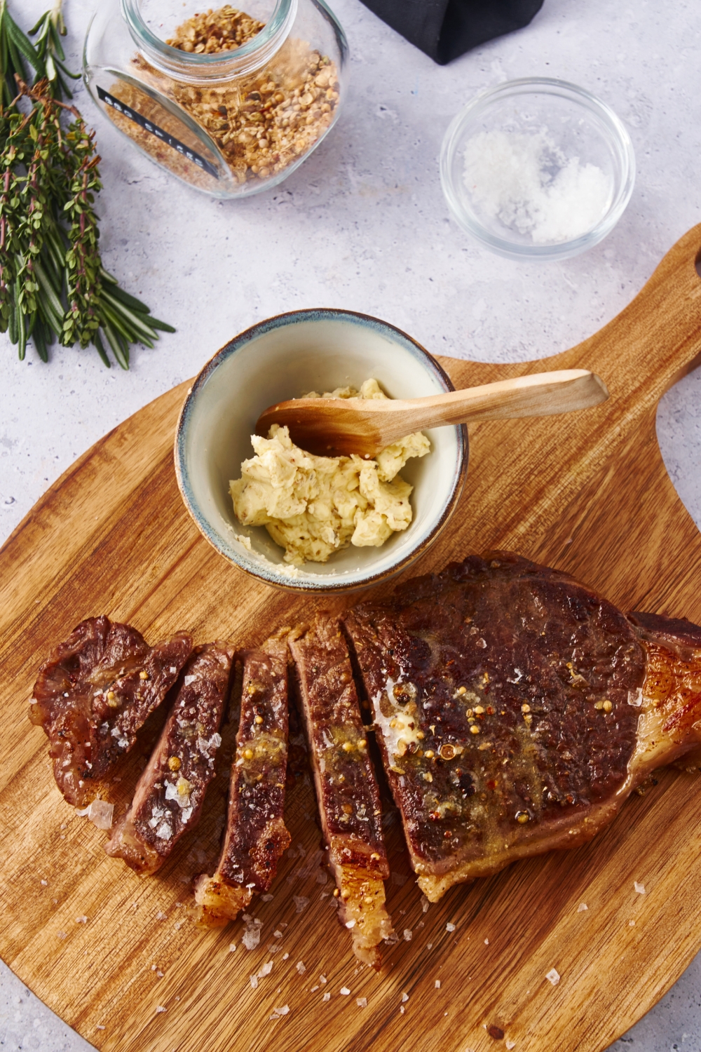 A cooked steak on a wood cutting board. Half of the steak has been sliced into thin slices and there is a bowl of compound butter on the board with a wood spoon in the bowl.