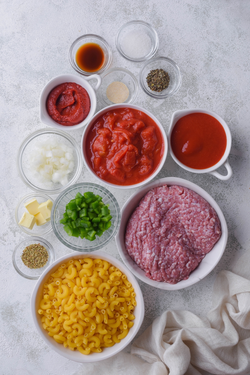 An assortment of ingredients including tomato sauce, tomato paste, raw ground beef, dried elbow macaroni, diced bell peppers, diced onion, spices, and butter.