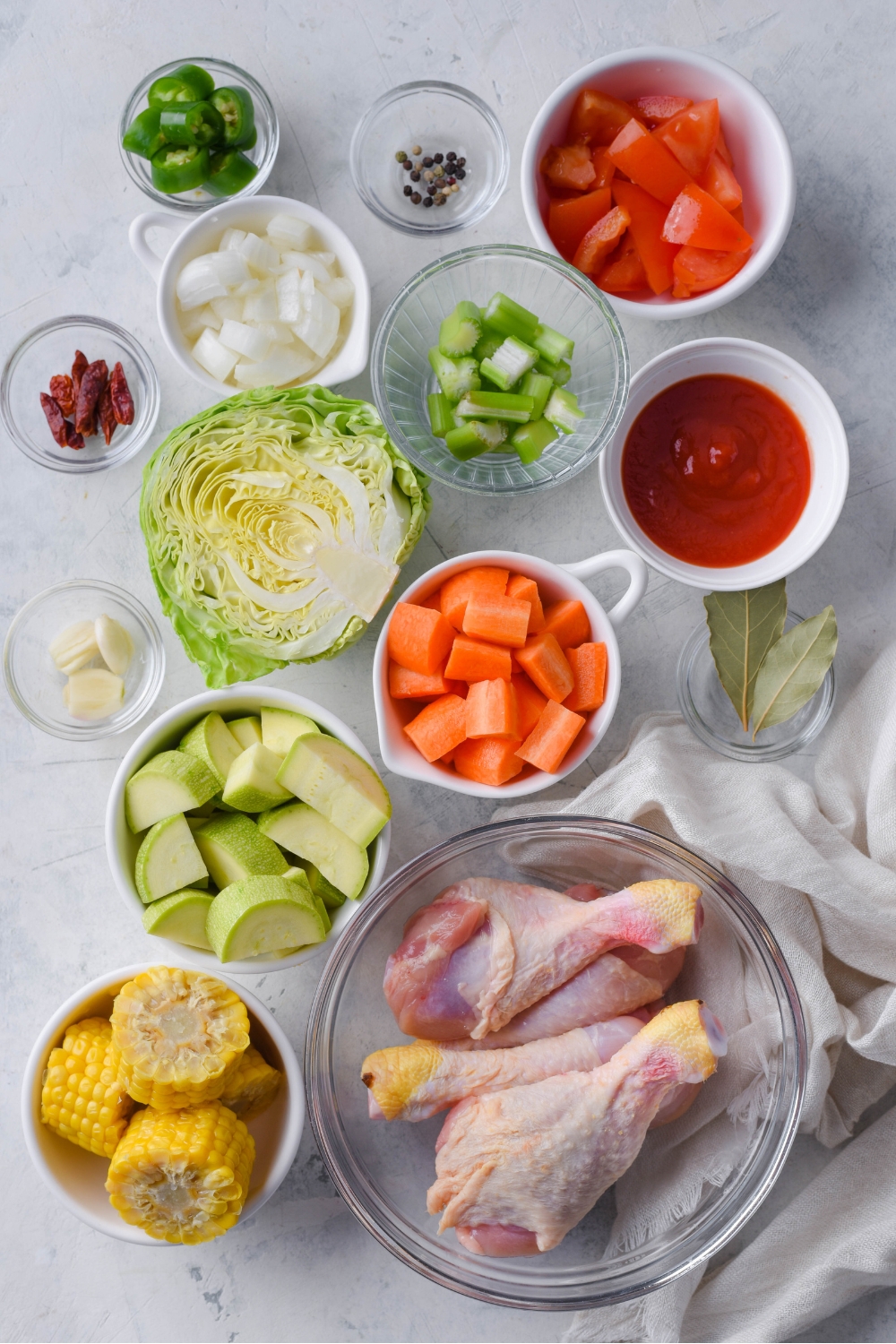 An assortment of ingredients including bowls of tomato sauce, celery, carrots, zucchini, corn, raw chicken, onion, and peppers.