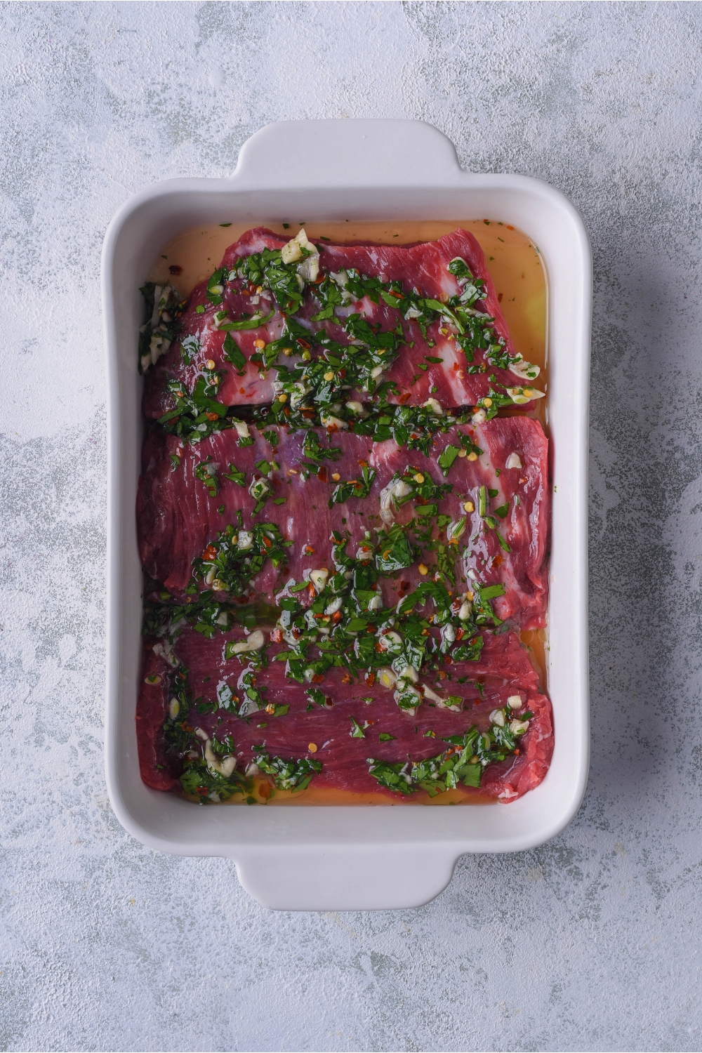 A white baking dish filled with bavette steaks marinating in a blend of green herbs.
