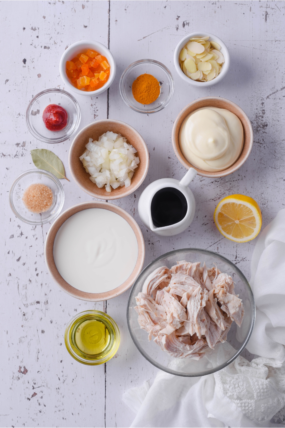 An assortment of ingredients including bowls of mayonnaise, creme fraiche, cooked chicken, oil, spices, and diced apricots.