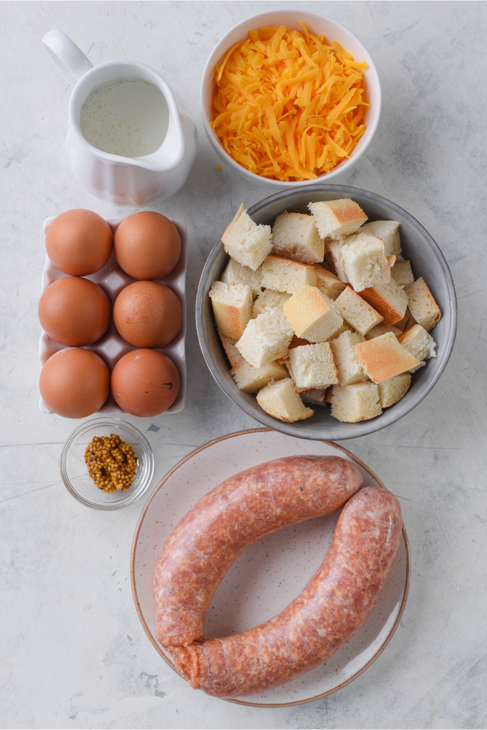 An assortment of ingredients including six eggs, a bowl of shredded cheese, a bowl of bread cubes, a plate of raw sausage, a container of milk, and a bowl of spices.
