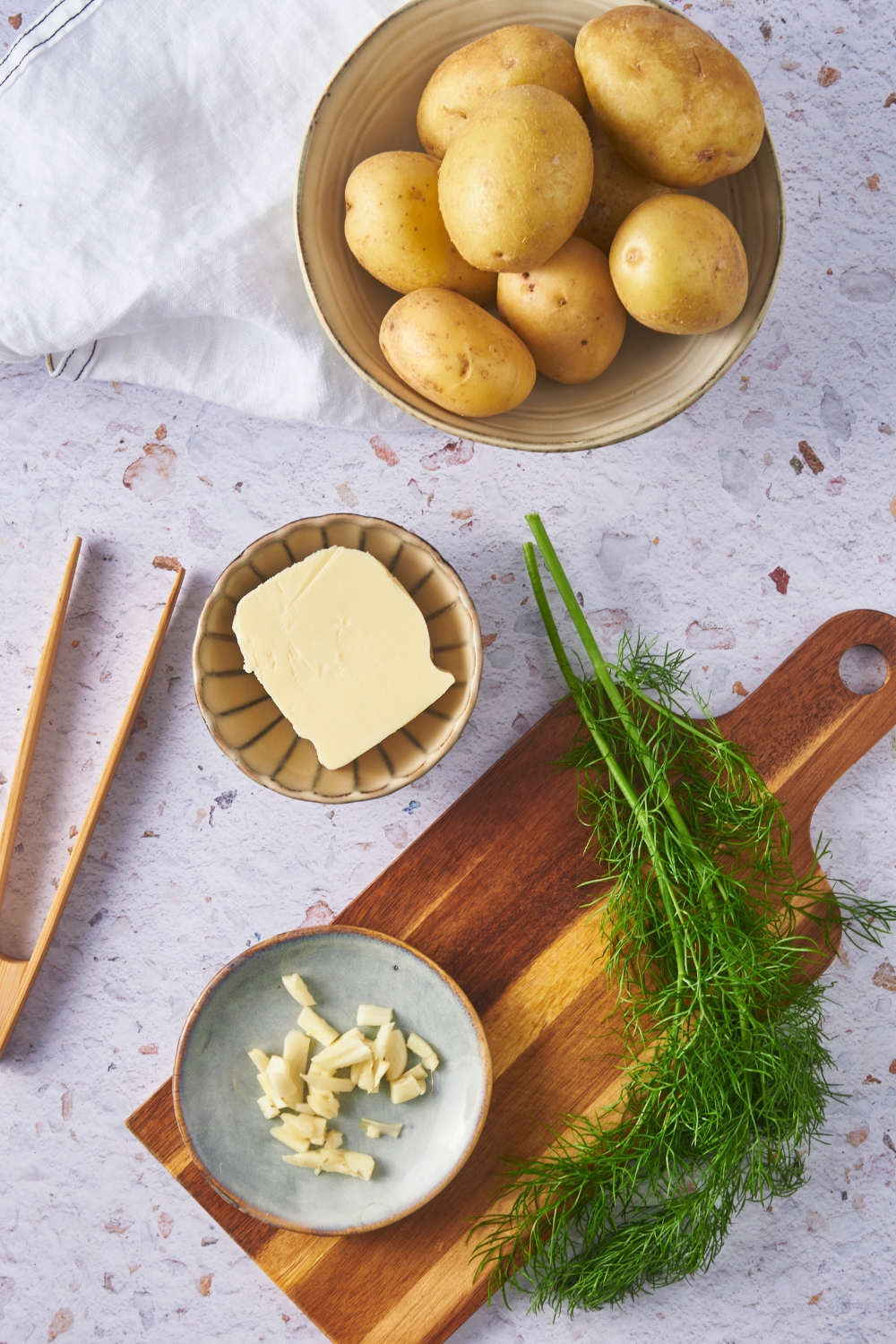 Overhead view of an assortment of ingredients including a bowl of potatoes, a bowl of butter, and a cutting board with fresh dill and a plate of sliced garlic on it.