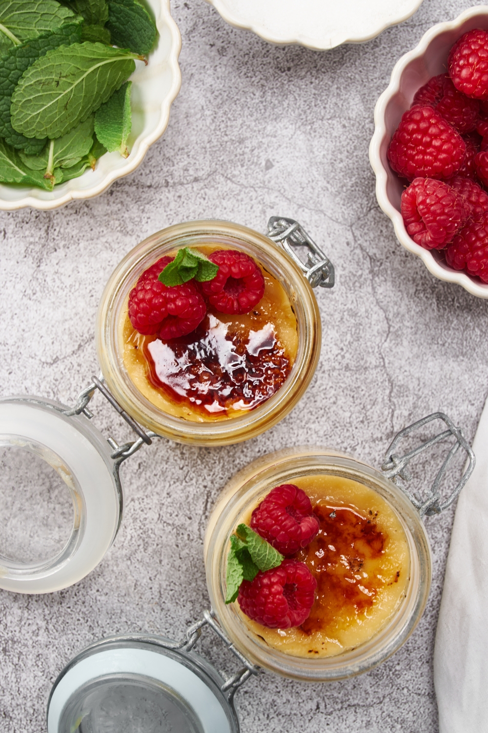 Overhead view of two creme brûlée desserts in jars, both next to bowls of fresh mint leaves and fresh raspberries.