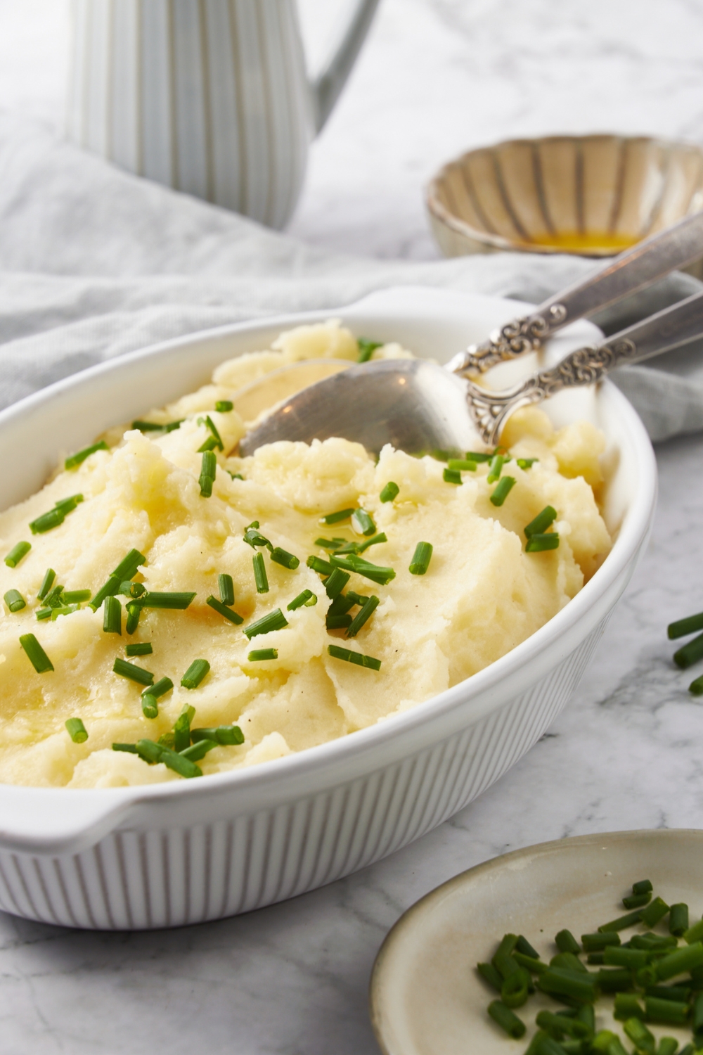 A white baking dish filled with mashed potatoes, two serving spoons, and fresh chives garnished on top.