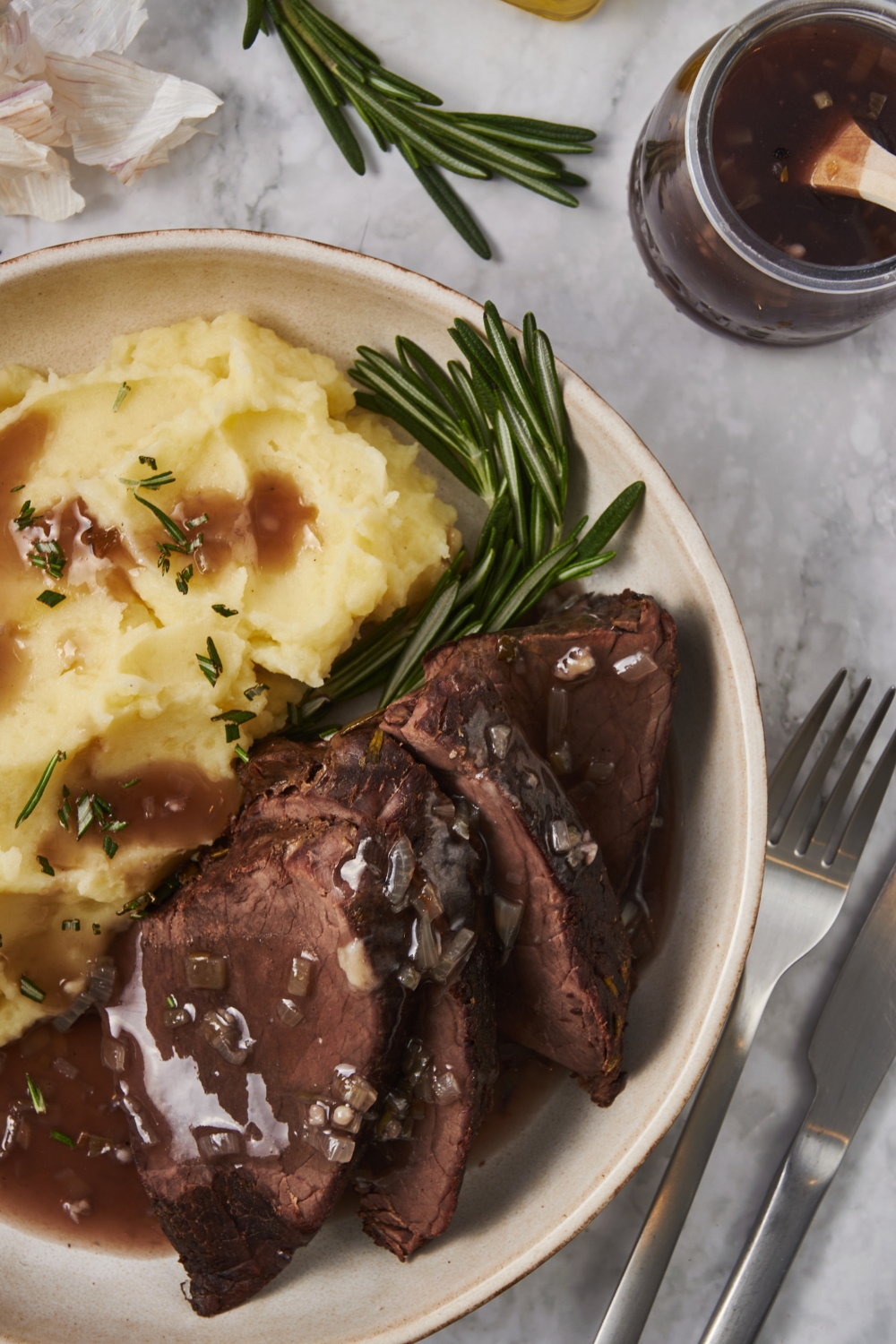 Close up of a plate of sliced chuck roast covered in a gravy with a fresh sprig of rosemary and a pile of mashed potatoes garnished with fresh herbs and some gravy poured over top of the potatoes.