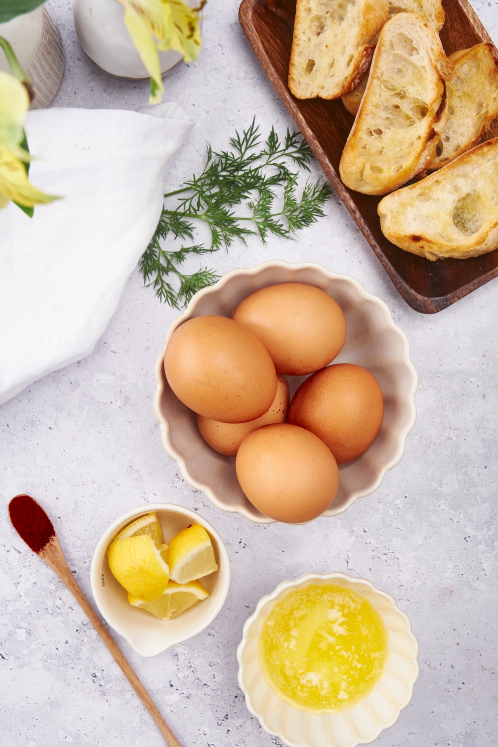 An assortment of ingredients including bowls of eggs, butter, lemon slices, a plate of toast, a sprig of dill, and a small spoon of cayenne pepper.