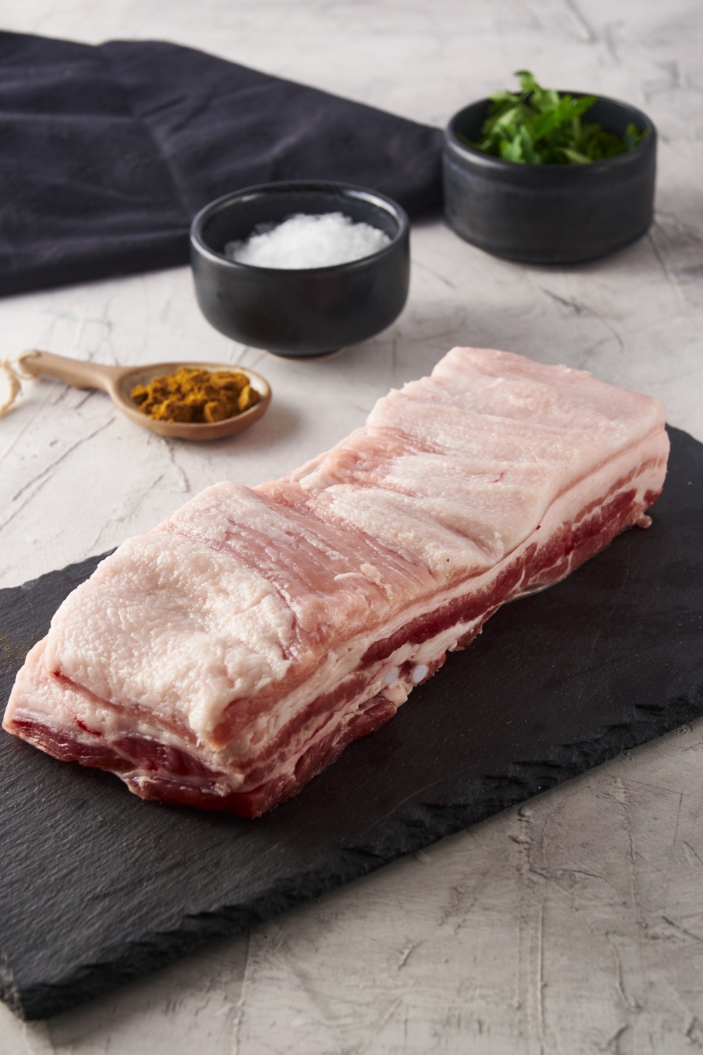Raw pork belly on black slate with seasonings in the background.