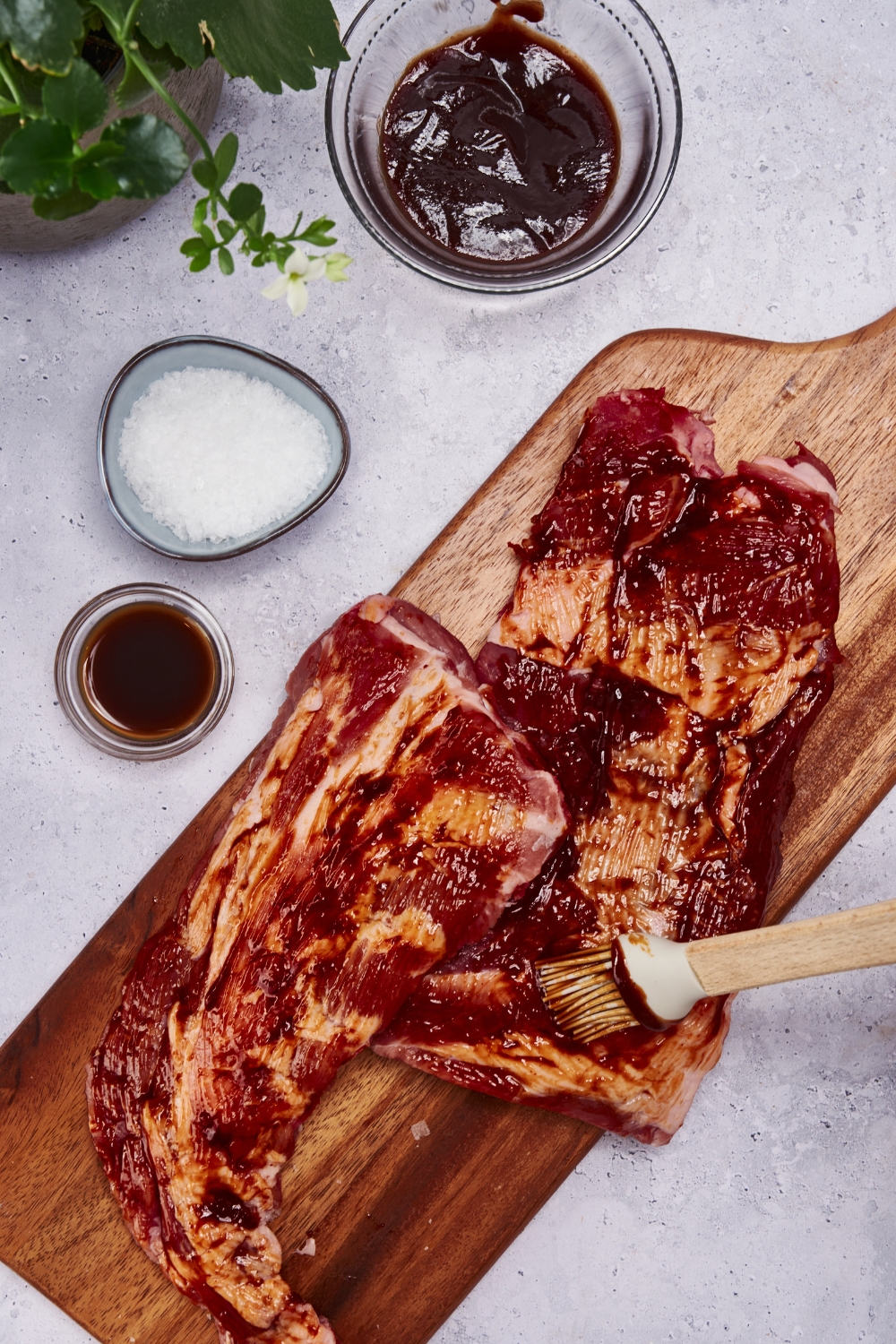 Two raw racks of ribs on a wood cutting board being covered in barbecue sauce with a silicone basting brush.