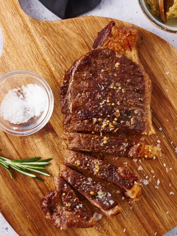 Overhead view of a cooked and sliced steak seasoned with salt. The steak is on a wooden board along with a bowl of salt.