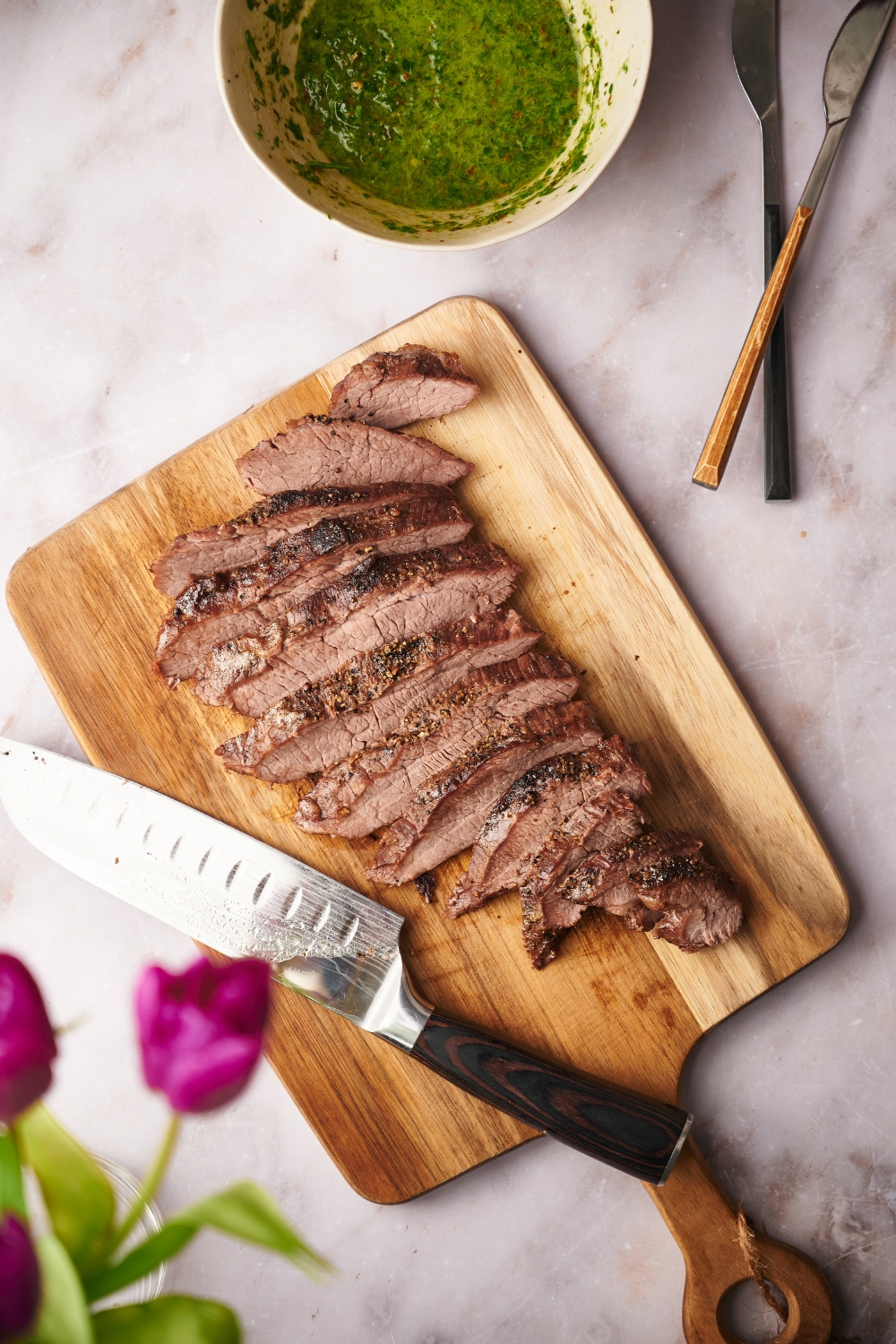 A cooked tri-tip steak that has been sliced into equal-sized thin slices. The steak is on a wooden board and there is a knife on the board along with a bowl of chimichurri sauce next to it.