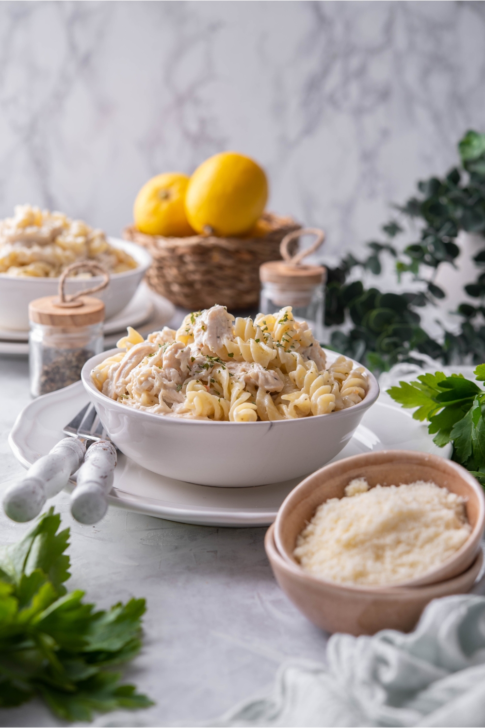 A bowl of cooked chicken and pasta in a cream sauce garnished with fresh herbs. The bowl is on a white plate with two forks on the plate and it is surrounded by ingredients including parsley, grated parmesan cheese, and a bowl of lemons.
