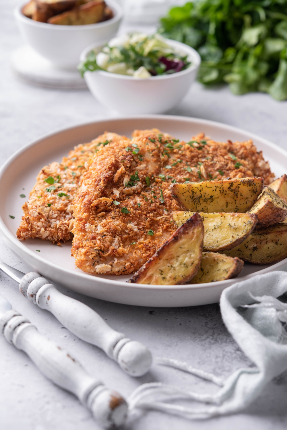 A plate of seasoned baked chicken cutlets garnished with fresh herbs and served with a side of potato wedges. There's a set of silverware next to the plate.