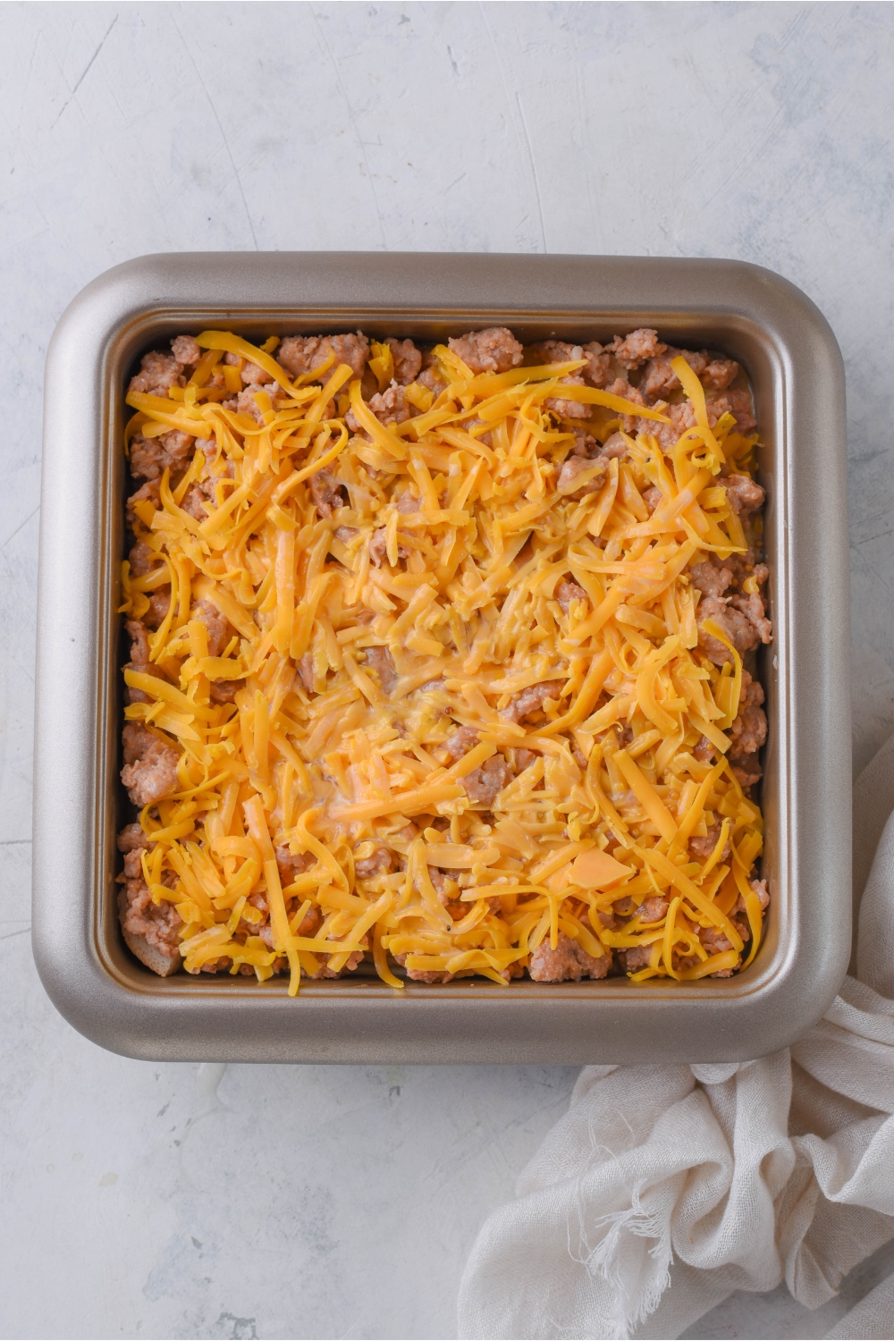 A square baking dish filled with cooked sausage, bread cubes, an egg mixture, and a layer of shredded cheese covering the dish.