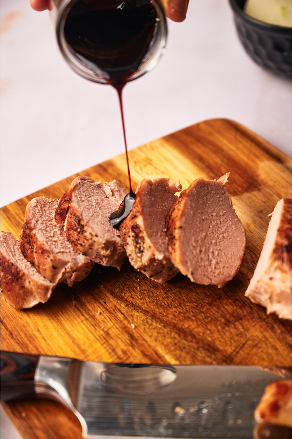 Close up of sliced and cooked pork tenderloin on a wood cutting board with a hand pouring a brown sauce over top the pork.