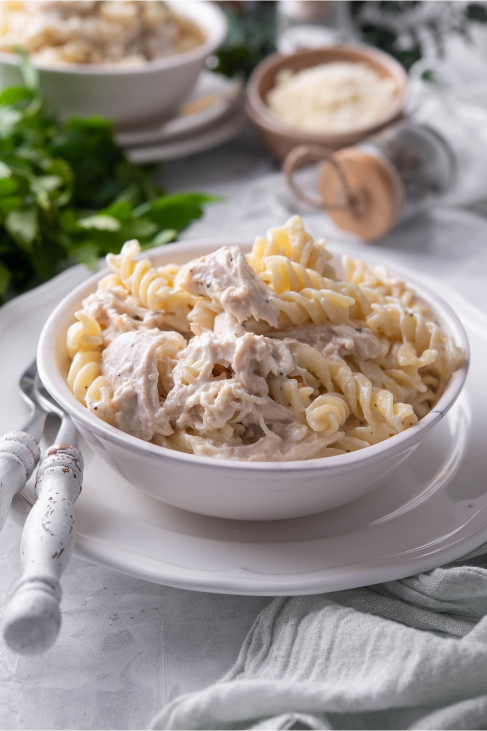 A bowl of pasta and chicken in a cream sauce atop a white plate with two forks next to the bowl of chicken and pasta.