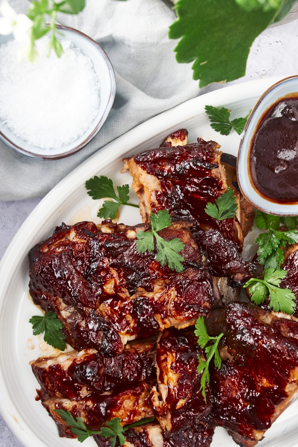 Overhead view of cooked ribs covered in barbecue sauce piled on top of each other on a white plate, with a side of barbecue sauce on the plate and fresh parsley garnished on top of the ribs.