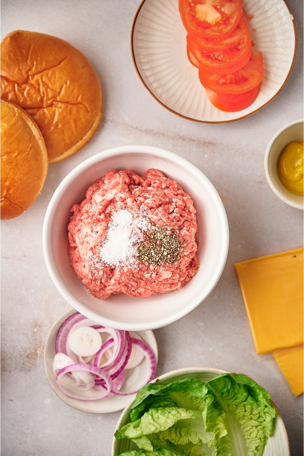 Overhead view of a bowl of raw ground beef with salt and pepper sprinkled on top. The bowl of beef is surrounded by ingredients for making cheeseburgers.