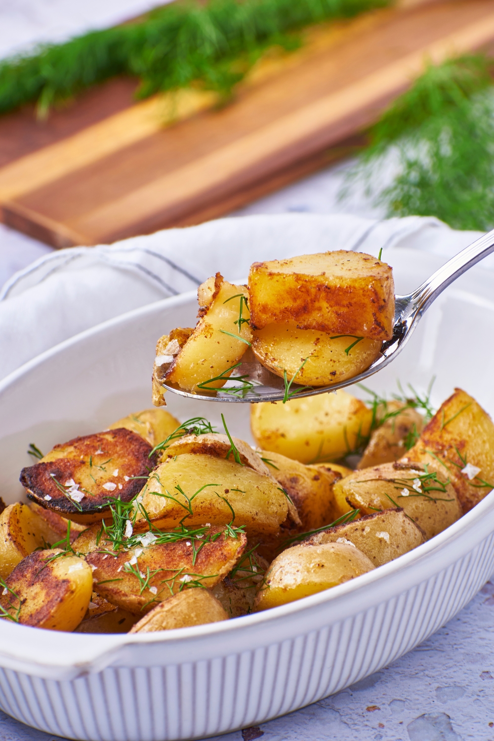 A dish of cooked potatoes seasoned with salt and fresh dill and a spoonful of potatoes being scooped out of the dish.