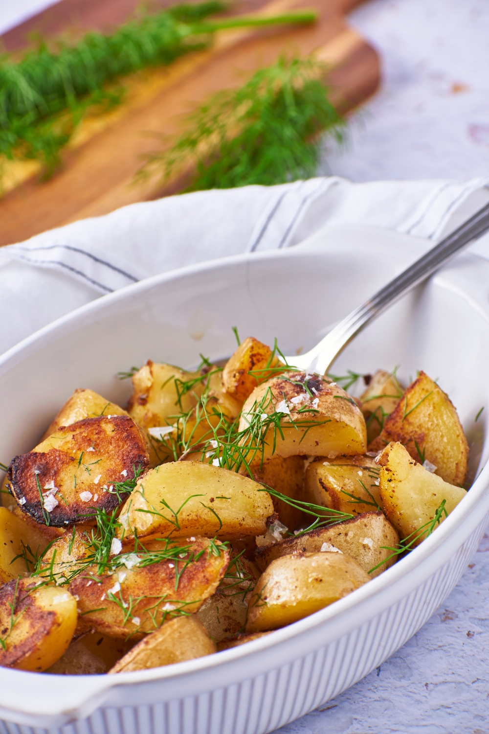 A white serving dish filled with cooked potatoes covered in sea salt and fresh dill. There is a serving spoon in the dish.