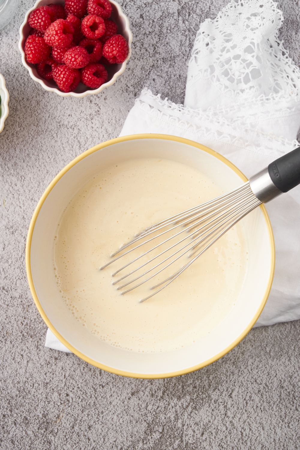 A white mixing bowl with custard and a whisk in it. A bowl of raspberries is next to the custard.